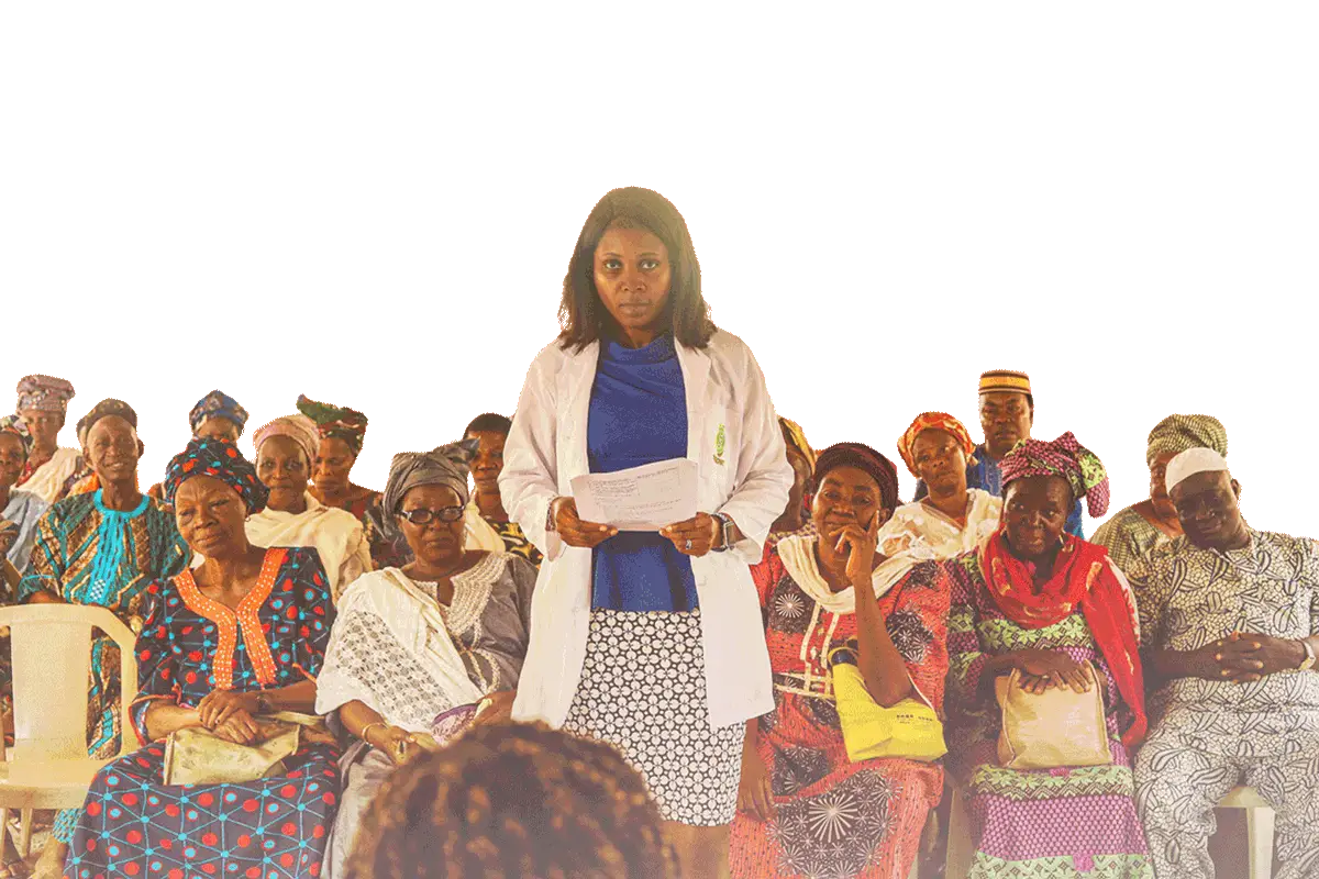 A doctor in Africa wearing a white coat and face mask stands holding papers, addressing a group of locals waiting to receive medical treatment.
