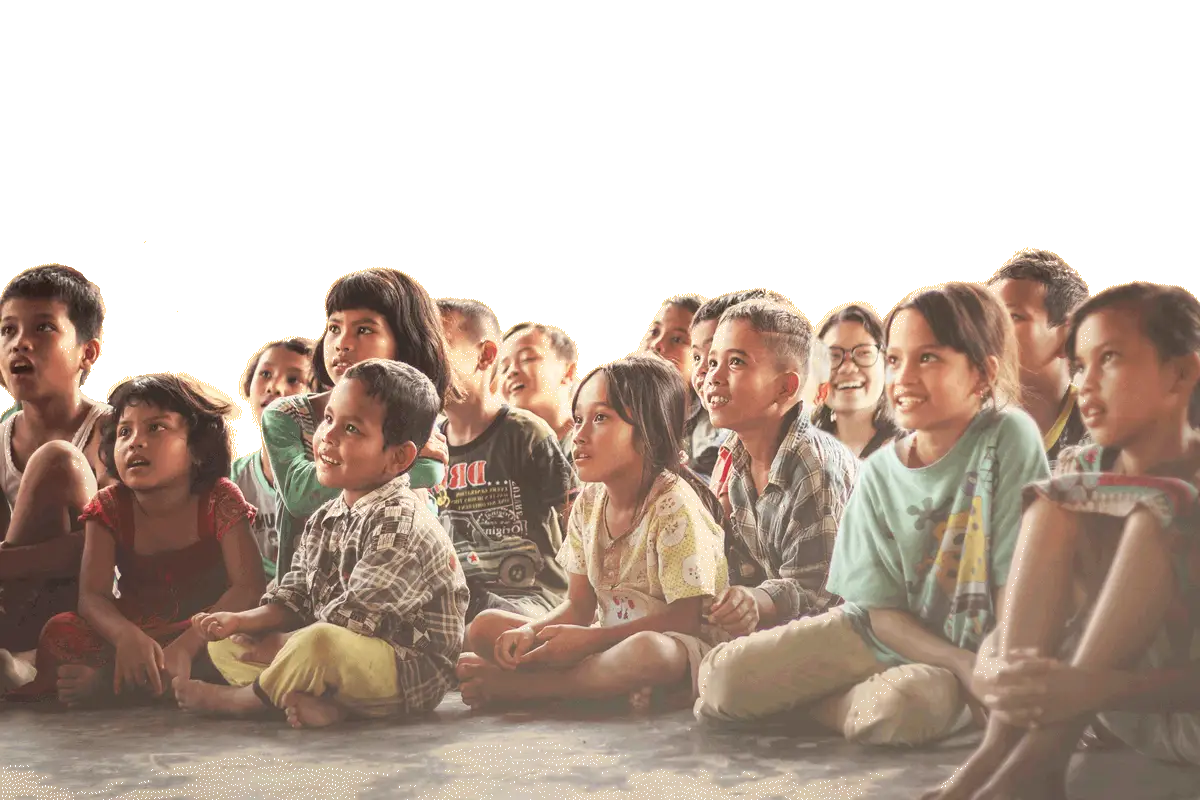 A group of children sitting closely together on the floor of a classroom, smiling and looking ahead, against a plain yellow background.